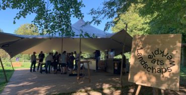 Een groep mensen onder een tentafdak met een bord aan de rechterkant.