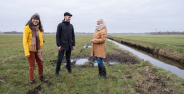 Foto van drie mensen in een weiland. Links een vrouw, in het midden een man en rechts nog een vrouw. Daarnaast een beekje.