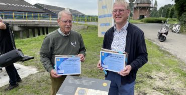 Twee mannen met een blauw certificaat in hun handen, poserend voor de foto.