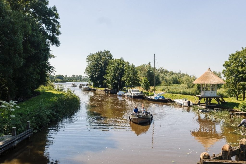 Landschapsbeeld met een slootje in het midden. Er vaart een bootje en er liggen bootjes aangemeerd. Aan weerszijden is groene natuur, met gras en bomen.