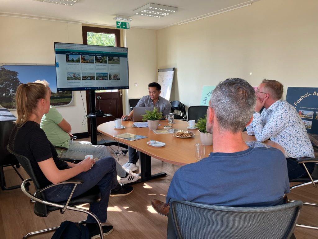 Een groep van 6 mensen aan tafel, waarbij een een presentatie geeft via een groot scherm