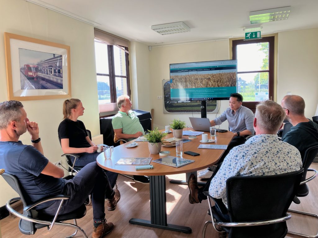 Een groep van 6 mensen aan tafel, waarbij een een presentatie geeft via een groot scherm