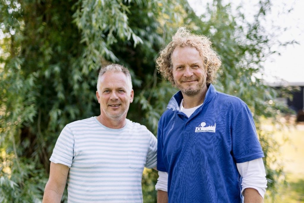 Twee mannen poseren voor de camera met natuur op de achtergrond.