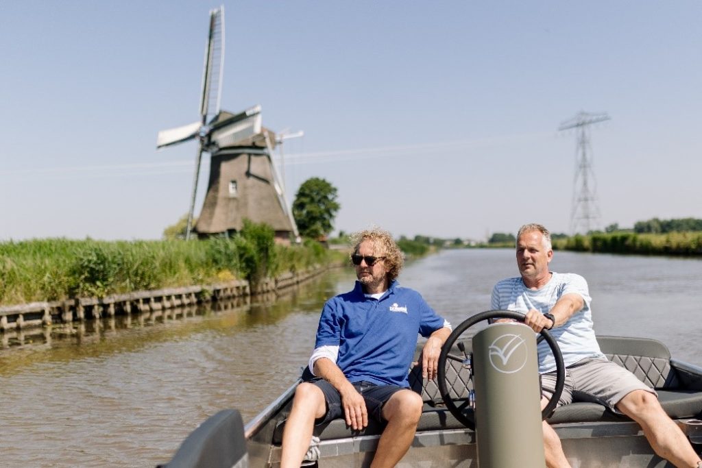 Twee mannen op een bootje, waarbij een het stuur vasthoudt. Op de achtergrond is water, gras en een molen te zien.