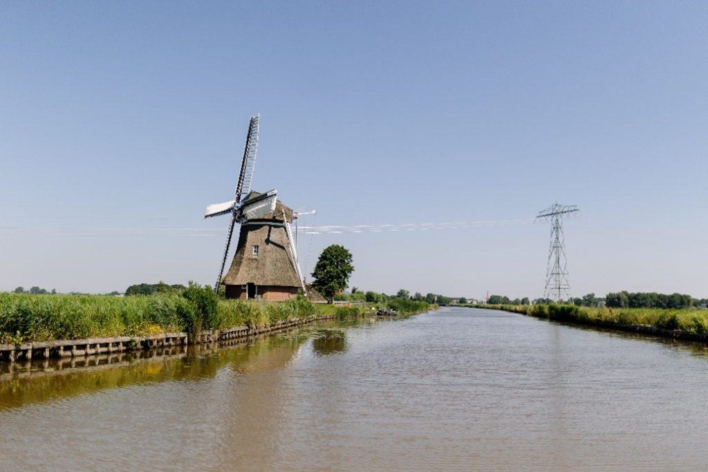 Een rustig beeld van water, met aan weerszijde hoog gras en links een molen. 