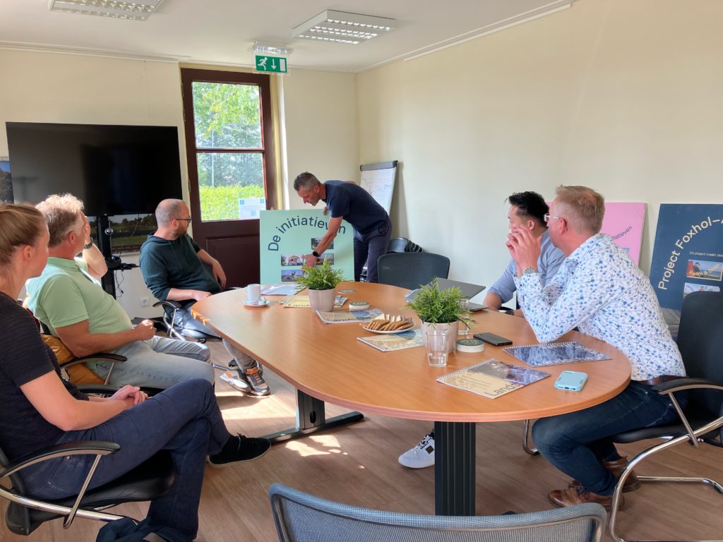 Een groep van 5 mensen aan tafel en een iemand geeft een presentatie bij een opgemaakt bord 