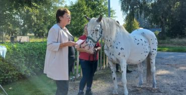Een vrouw links en een paard rechts. De vrouw spreekt voor een aantal mensen.