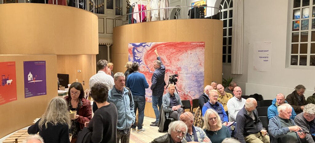 Een groep mensen in een zaal. Rechts zitten, voornamelijk oudere mensen, op stoelen. Op de achtergrond staan een aantal mensen voor posters aan muren.