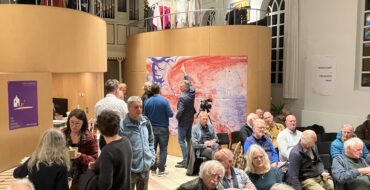 Een groep mensen in een zaal. Rechts zitten, voornamelijk oudere mensen, op stoelen. Op de achtergrond staan een aantal mensen voor posters aan muren.