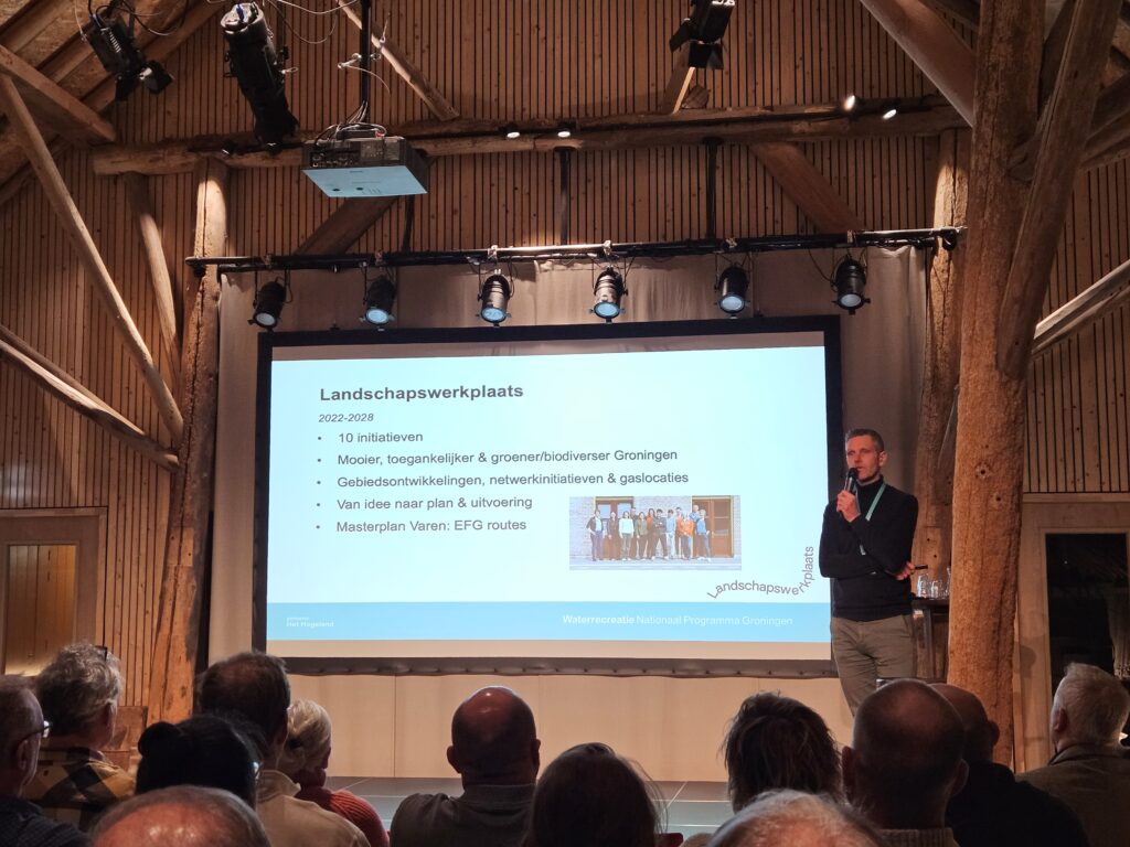 Man staat rechts van een groot scherm met een microfoon in zijn hand. Hij praat tegen het publiek in de zaal.