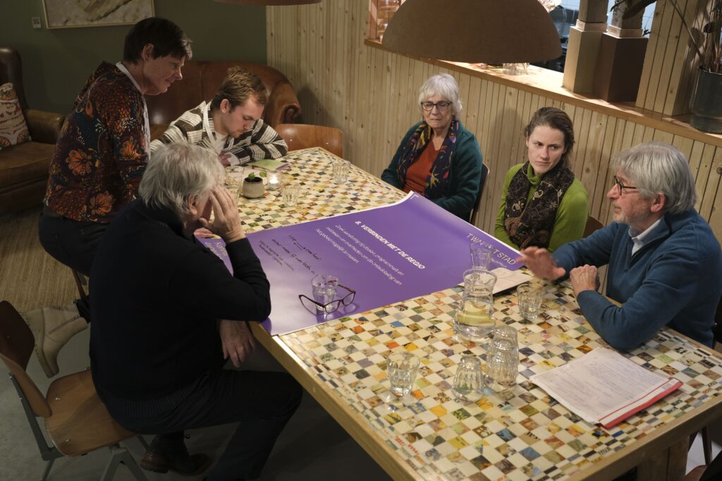 Mensen zitten in een groepje rond een tafel. Hierop ligt een paars papier. Mensen zijn in gesprek.
