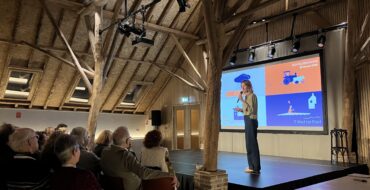 Iemand op een podium voor een scherm. Publiek kijkt vanaf de zaal.